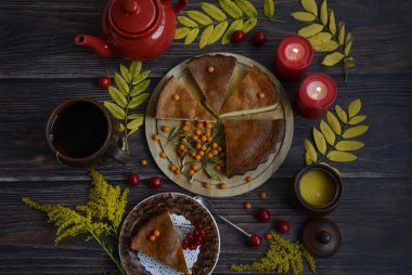 Tahta tabakta ev yapımı turta, kırmızı çaydanlık, seramik kupa, mumlar, deniz dikenleri ve dikenli üzümler, koyu ahşap arka planda sarı sonbahar yaprakları.
