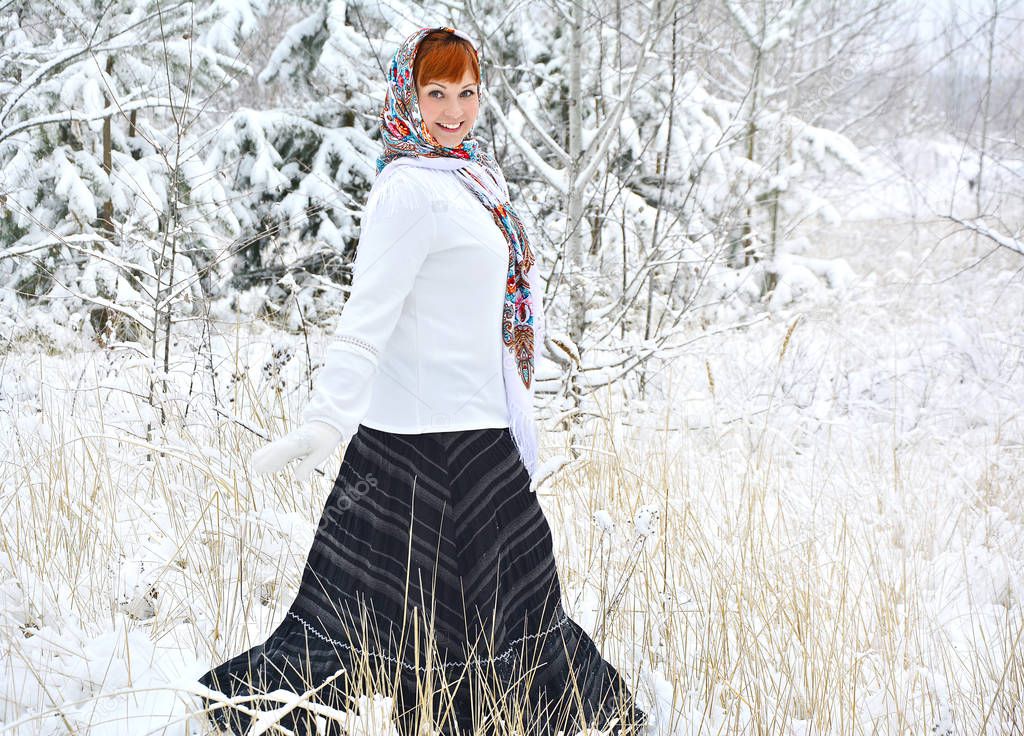 A red-haired smiling girl in a bright chalet and white clothes is walking through a snowy forest