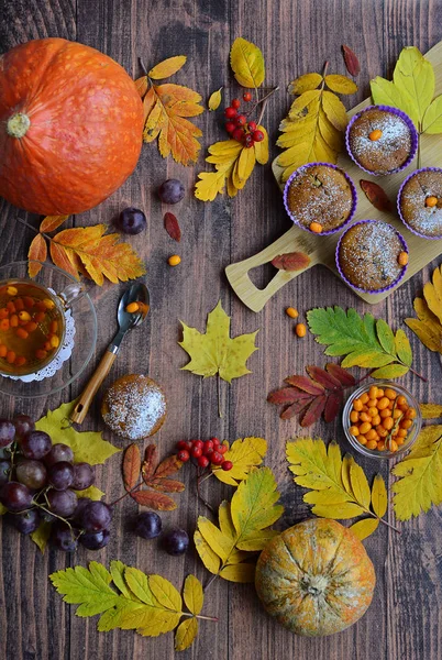 Composizione autunnale di fogliame colorato, bacche, cupcake di zucca, uva, tè con olivello spinoso su uno sfondo di legno — Foto Stock