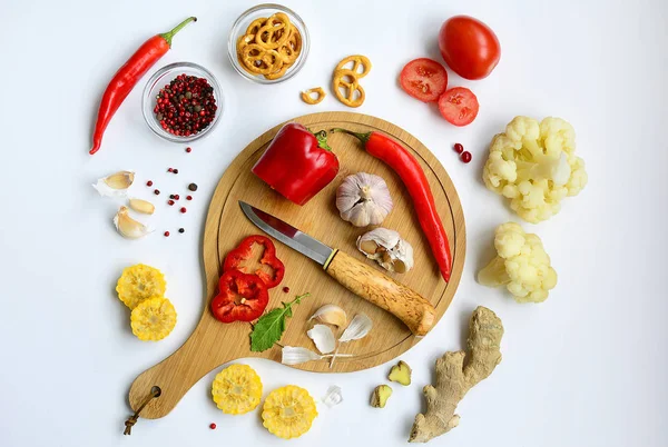 Κυκλική διάταξη από κόκκινες πιπεριές και ντομάτες, κίτρινο καλαμπόκι, σκόρδο, κουνουπίδι, τζίντζερ, κράκερ, ξύλο κοπής με μαχαίρι σε λευκό φόντο — Φωτογραφία Αρχείου