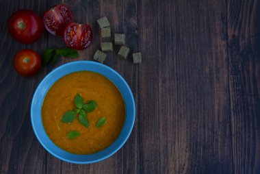 Appetizing soup puree in a blue deep plate, tomatoes and slices of dried bread on a dark wooden background
