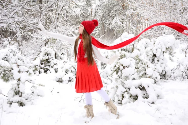 Mladá dívka v bílém svetru a červeném pleteném klobouku a létajícím šátku se dívá stranou a mává rukou na pozadí zasněženého zimního lesa — Stock fotografie
