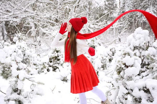 Menina bonita nova em um chapéu vermelho brilhante, luvas e um lenço desviam os olhos em uma floresta conífera nevada — Fotografia de Stock