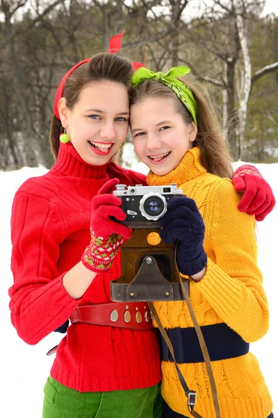 60 years style. Two girls in red and yellow sweaters playfully smile at the lens and are holding an old camera. Vertical