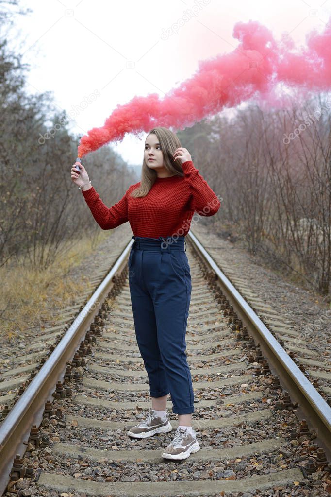 A young girl in a red sweater stands on the rails with red smoke in her hands and looks dreamily into the distance. Bright fashion concept. Vertical