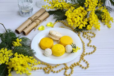 Hindistan cevizli kurabiyelerin ve beyaz bir tabakta sarı badem kurabiyelerinin üst görüntüsü, mimoza dalları, el işleri ruloları ve hafif ahşap arka planda altın boncuklar. Durgun hayat.