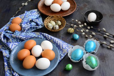 Mavi ve kahverengi tabakta boyanmamış yumurtalar ve koyu mavi masa arkaplanındaki parlak mavi ve yeşil paskalya yumurtaları. Paskalya gelenekleri. Kapat.