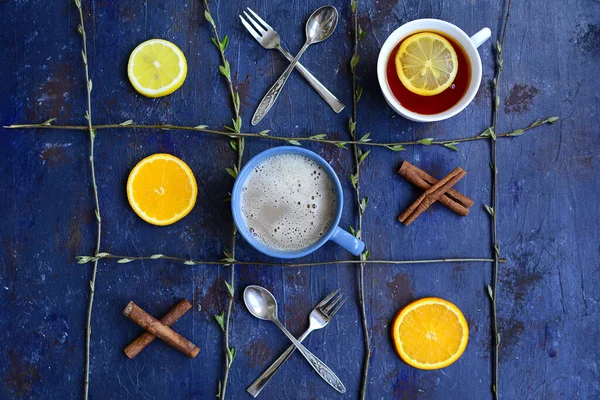Cups with tea and coffee, circles of lemon and orange, cinnamon sticks, forks and spoons in the shape of a tic-tac-toe game. View from above.