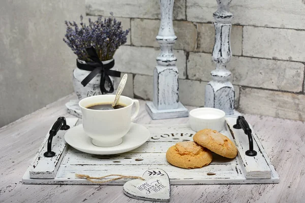 White cup of coffee and homemade cookies on a wooden tray, dry lavender in a vase and candlesticks in the style of shabby on a rustic table. Close-up