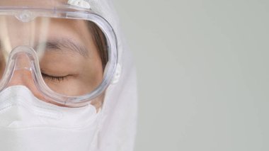 Koruyucu tehlikeli madde giysileri içinde, yüz maskesi ve fotokopi alanı olan gözlük takan Asyalı doktorun gözlerini kapat.