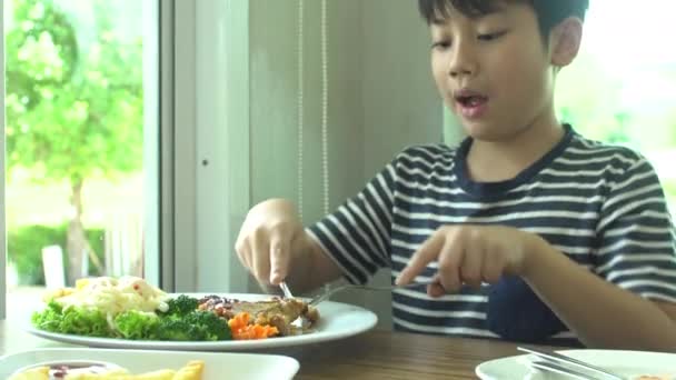 Kleiner asiatischer Junge isst Steak mit Gemüsesalat im Restaurant mit einem Lächeln im Gesicht — Stockvideo