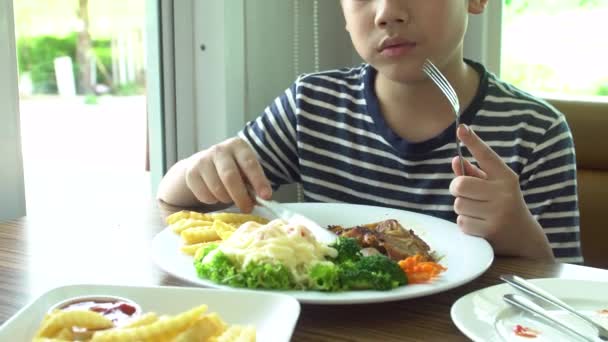 Азиатский мальчик ест стейк с овощным салатом в ресторане с улыбкой на лице — стоковое видео