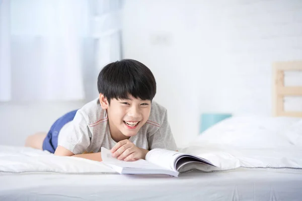 Ásia menino resto no o cama e leitura livro com sorriso rosto — Fotografia de Stock