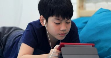 Küçük Asyalı çocuk dinlenme mavi yatakta kulak telefon ile tablet bilgisayar kullanarak .