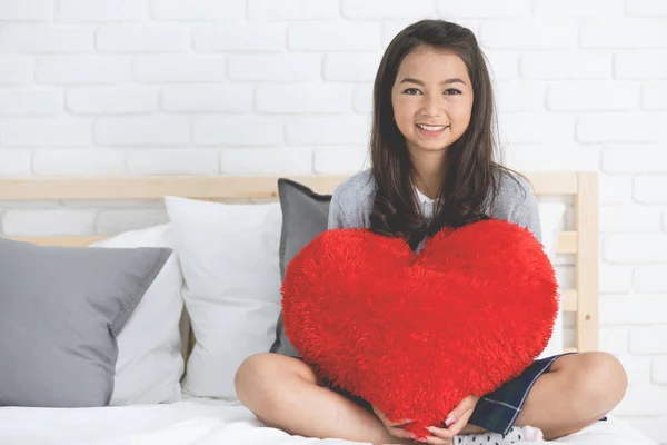 Porträtt av asiatiska söt flicka sitter på sängen med stor röd kudde s — Stockfoto