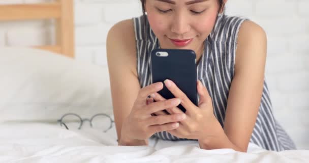 Mulher asiática bonito sorrindo com seu telefone celular na cama falando e mensagens de texto  . — Vídeo de Stock