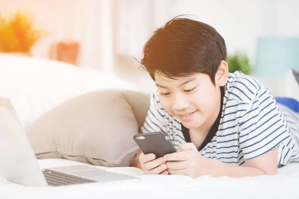 Lindo asiático chico resto en cama y usando inteligente celular con sonrisa —  Fotos de Stock