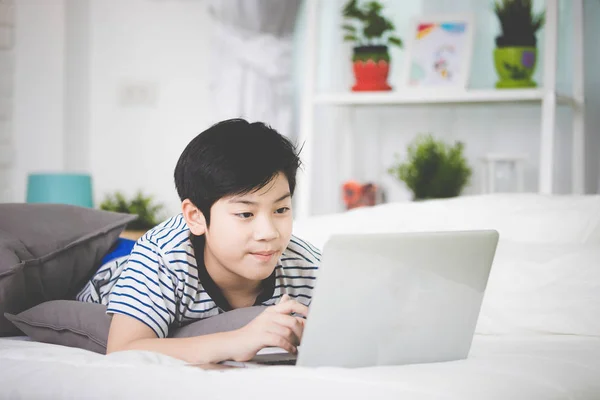 Sød asiatisk dreng hvile på sengen og skrive bærbar computer . - Stock-foto