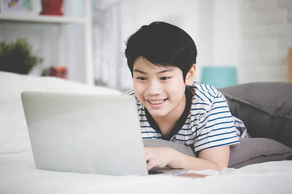 Lindo asiático chico resto en cama y mecanografía laptop ordenador . — Foto de Stock