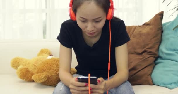 美しいアジアの女性音楽イヤホンを聞き 自宅のソファでスマート フォンを使用してリラックスしたライフ スタイル — ストック動画