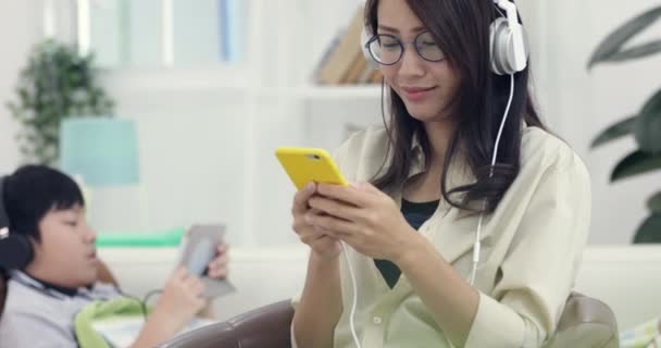 年轻的亚洲妇女放松和听音乐与男孩玩平板电脑在客厅里 — 图库视频影像