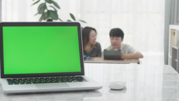 リビングルームの机の上に緑のクロマキー画面が立っているノートパソコン 背景の母親と息子がタブレットPcで一緒に見ている — ストック動画