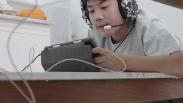 アジアの少年タブレットコンピュータで友人とオンラインゲームをプレイ 若い男性は興奮してマイクでヘッドフォンを通して話しています — ストック動画