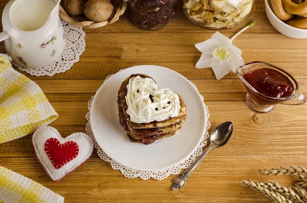Buñuelos, panqueques en forma de corazones con mermelada y crema batida sobre un fondo de madera clara con un corazón rojo, un desayuno romántico para el Día de San Valentín. Día de las Madres. Cocinar — Foto de Stock