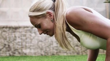 Beyaz tişörtlü güzel sarışın bir kız temiz havadaki yeşil çimlerde spor egzersizleri yapıyor. Arka planda beyaz ve gri bir duvar var..