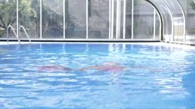 Pembe mayo giymiş iki sarışın mavi su havuzunda yüzüyor..