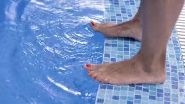 İki çift kadın bacağı havuzun kenarında duruyor ve mavi berrak su ile parmaklarını hareket ettiriyor..