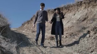 Siyah şapkalı genç adam ve kadın kumlu tepeden iner, güneşli bahar gününde birbirlerinin ellerini tutarlar.