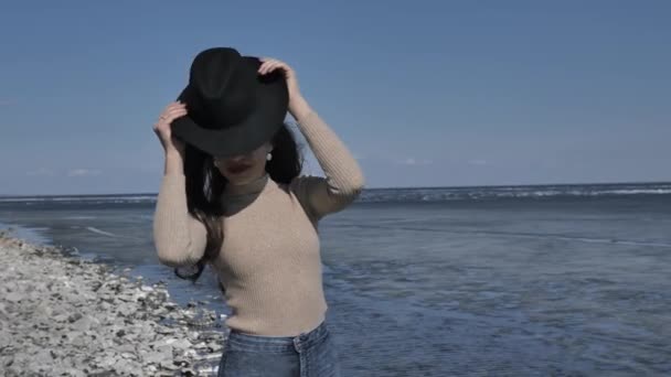 年轻美丽的姑娘戴着黑色的帽子 看着相机 微笑着 背景是一条被冰覆盖的河流 春日阳光明媚 — 图库视频影像