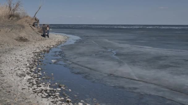 若いカップルは 川の岩の銀行に沿って 氷で覆われ 互いの手 晴れた春の日を保持歩いてください — ストック動画