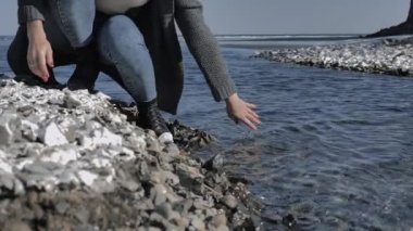 Young girl moisturizes her hand, dipping in clear water on a rocky shore, sunny day