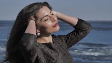 A young girl in a black dress strokes her hair, in the background the ocean, sunny day