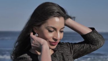 A young girl in a black dress strokes her hair, in the background the ocean, sunny day