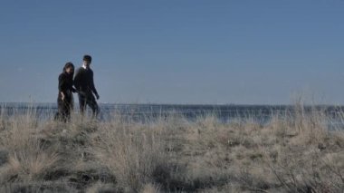 Young couple dressed in dark walks along the seashore, sunny day.