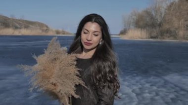 A young Caucasian girl in a black dress holds a cane bouquet in her hands, in the background a river covered with blue ice, a sunny day