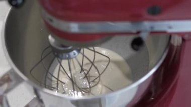 Bir kadın eli, kırmızı profesyonel bir mikseri beyaz sıvıyla metal bir kaba daldırır ve yakın plan olarak çalıştırır..