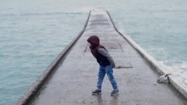 Bir çocuk beton bir binadan mavi deniz suyuna taş atıyor..