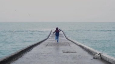 Çocuk denizdeki beton bir yapıda koşuyor ve uzaktaki martıları dağıtıyor. Dalgalar taş sahile vuruyor..