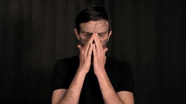 Siyah elbiseli beyaz bir adam siyah bir arkaplanda, kameranın önünde yüzüne kozmetik kil sürüyor..