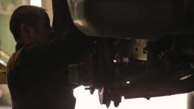 Asyalı bir adam atölyedeki asansörde kaldırılan eski bir arabanın şasesini tamir ediyor..