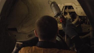 Asyalı bir adam atölyedeki asansörde kaldırılan eski bir arabanın şasesini tamir ediyor..