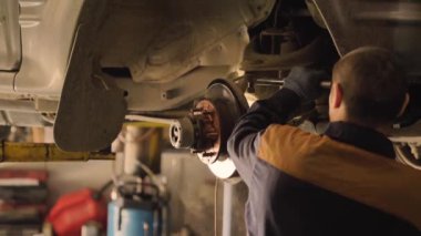 Asyalı bir adam atölyedeki asansörde kaldırılan eski bir arabanın şasesini tamir ediyor..