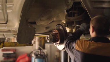 Asyalı bir adam atölyedeki asansörde kaldırılan eski bir arabanın şasesini tamir ediyor..