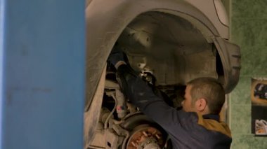 Asyalı bir adam atölyedeki asansörde kaldırılan eski bir arabanın şasesini tamir ediyor..