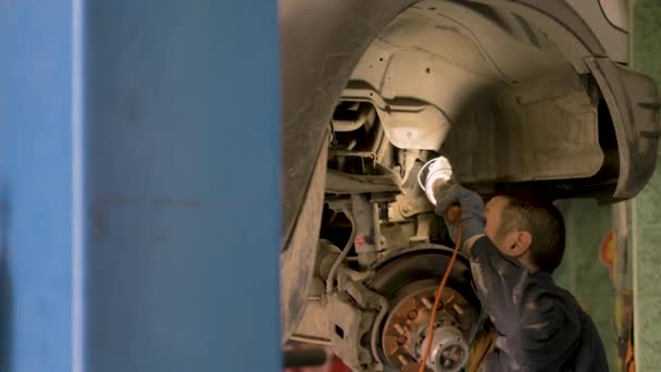 ワークショップでリフトで持ち上げられた古い車のシャーシを修理しているアジアの男 クローズアップ — ストック動画