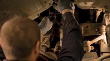 Asyalı bir adam atölyedeki asansörde kaldırılan eski bir arabanın şasesini tamir ediyor..